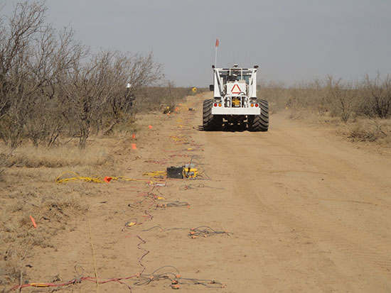 Color photo of seismic-source vehicle on dirt road in desert; geophones laid out along side of road, connected by wires.