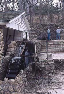 Stone trough with water from springs next to white wood-sided building; steep hillside in background covered with leaveless trees.